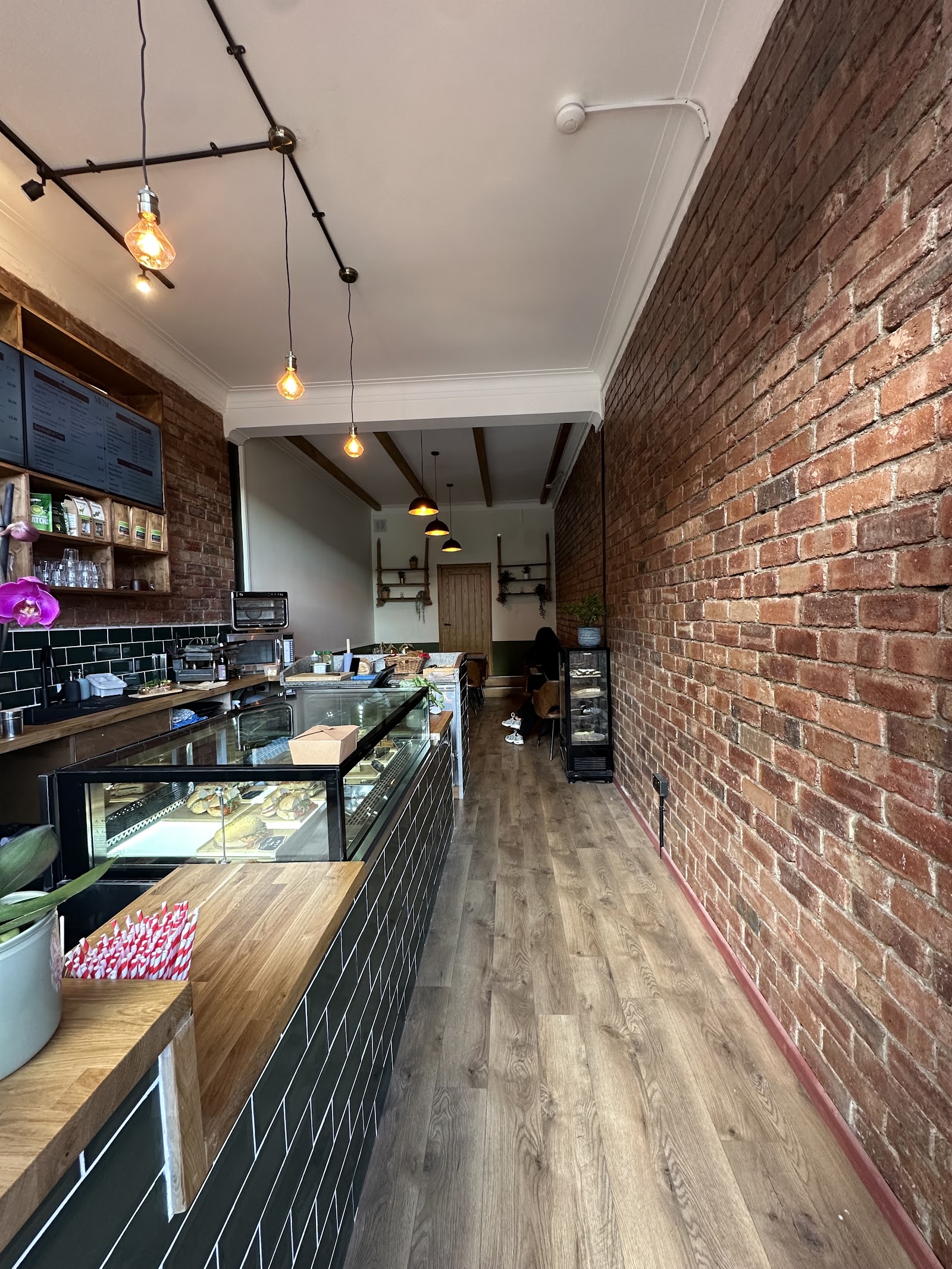 Inside Café Cor'stone: exposed brick wall, green tiled counter full of sandwiches, Edison bulb pendants